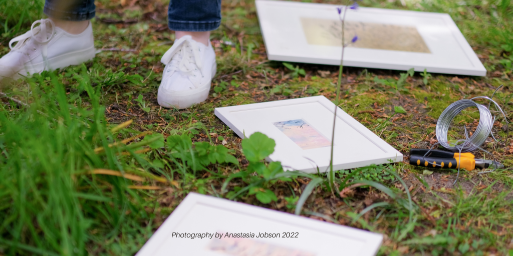 Artist Charlie Kirkham photographed by Anastasia Jobson wearing white sneakers next to three framed oil pastel and ink drawings in a woodland. Plyers and wire on display on ground