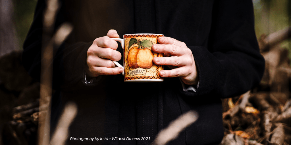 Charlie Kirkham photographed with a morning mug of coffee in an apricot mug, wearing a black winter coat in woodland. Photo by In Her Wildest Dreams
